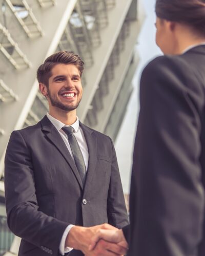 Handsome young businessman in classic suit is shaking hands with his colleague and smiling, standing outside the office building